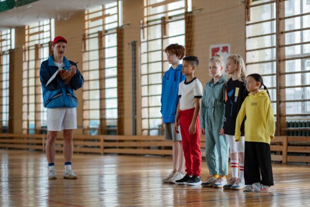 Grup de copii la un antrenament de sport într-o sală de sport școlară, alături de antrenor