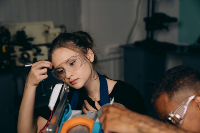 Elev cu salopetă de lucru care învață o meserie într-un atelier-școală, lângă un instructor